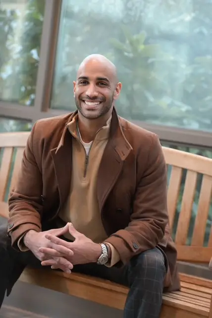 Jas Chahal seated on a wooden bench, smiling, in a brown coat